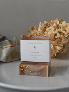 Bar of soap with 'Meadow Soap Co' packaging on a gray surface with dried flowers and a towel in the background.