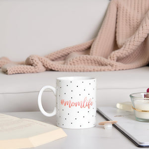 White mug with black polka dots and '#momlife' text on a table with a blanket and candle in the background